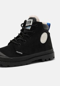 Bottines en suède noir dotées d'une semelle en caoutchouc, d'un laçage, d'un col en fourrure et d'un logo rond sur le côté. Tirette bleue à l'arrière.