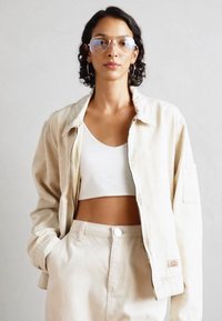 Chaqueta de denim clara con cuello, que presenta un cierre de cremallera y un bolsillo. Llevada sobre un top corto blanco y un conjunto a juego.
