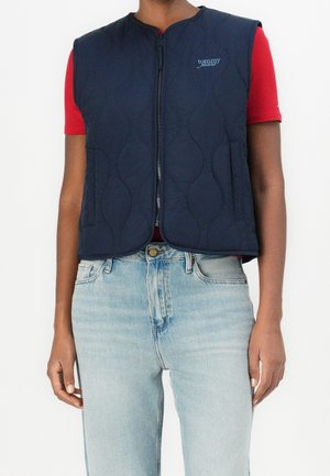 Osoba nosząca granatową pikowaną kamizelkę bez rękawów z zamkiem błyskawicznym z przodu i logo "Tommy Jeans" na czerwonej koszulce z krótkim rękawem oraz jasnoniebieskich dżinsach.