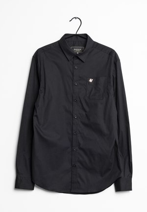 Chemise noire à manches longues, boutonnée, avec col et poche poitrine ornée d'un petit logo blanc brodé, suspendue à un cintre noir.