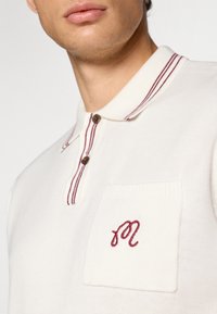 Beige polo skjorte med burgunderstripet krage, to-knapps placket og en brystlomme med brodert "M." Myk strikket tekstur.