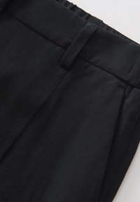 Détail d'un pantalon en tissu noir montrant la ceinture avec un dos élastique, une passants de ceinture et des plis à l'avant.
