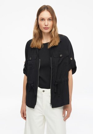 Frau mit schulterlangem Haar trägt eine schwarze kurzärmelige Reißverschlussjacke über einem schwarzen Top und weißen Hosen, steht vor weißem Hintergrund.
