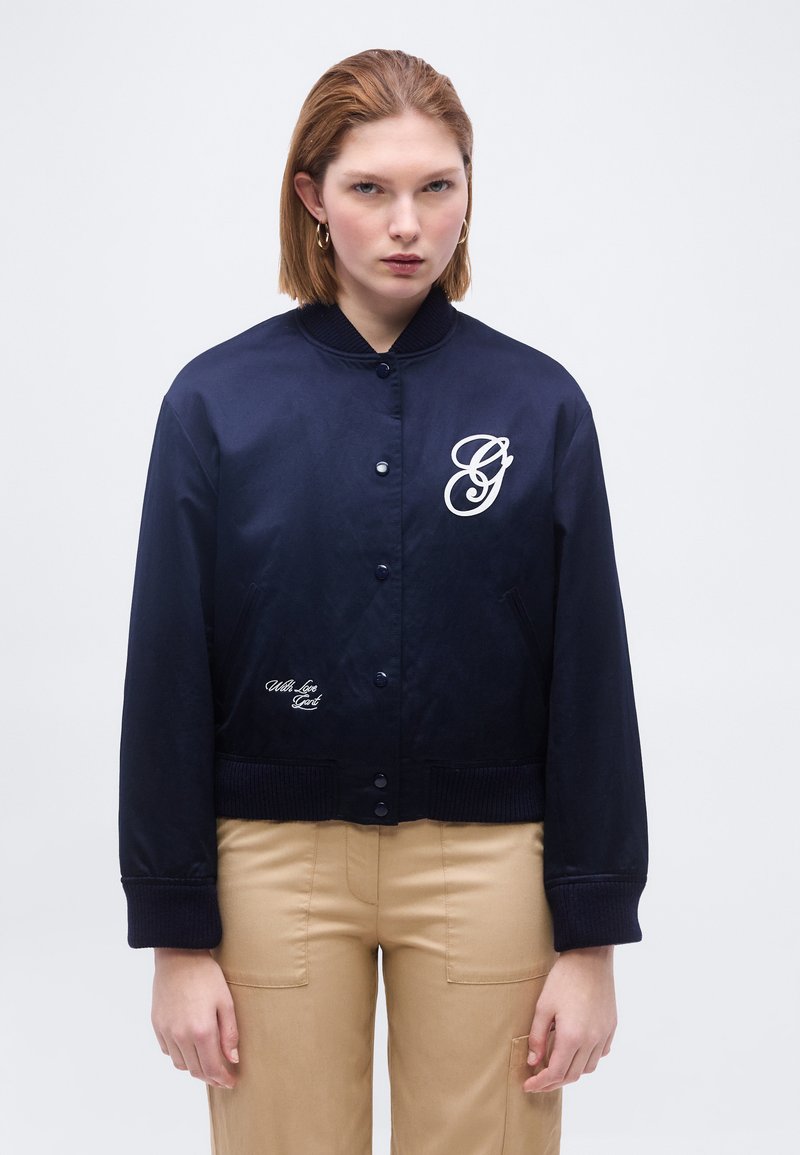 Jeune femme aux cheveux courts bruns, portant une veste bleu marine boutonnée avec un logo blanc et un pantalon beige, debout devant un fond uni.