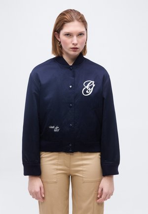 Ung kvinde med kort brunt hår iført en marineblå jakke med knapper og hvidt logo samt beige bukser, stående foran en ensfarvet baggrund.