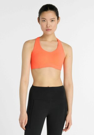 Kvinde iført en orange sports-bh og sorte høj-taljede leggings, stående mod en ensfarvet hvid baggrund.