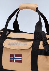 Sac de sport tan avec des anses noires, doté d'un logo, d'une poche avant et d'un écusson du drapeau norvégien. Tissu durable avec une texture lisse.