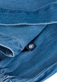 Denimstoff in Blautönen, mit einem gefalteten Design und einem kleinen Markenschild, das innen eingesteckt ist. Weiche Textur mit sichtbaren Nähten.