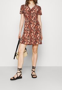 Robe florale à manches courtes, base noire, avec des fleurs rose-orange. Forme évasée, portée avec des sandales noires à lanières et un petit sac.