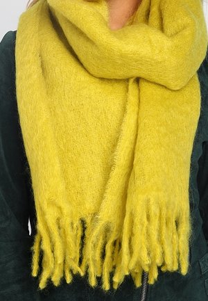 Écharpe jaune douce et duveteuse avec des extrémités à franges, fabriquée dans un matériau chaud, enroulée autour du cou, superposée à une veste verte.