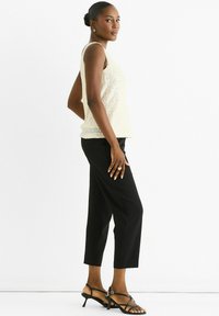 Woman standing sideways wearing a sleeveless cream sequin top, black cropped pants, black strappy heels, and gold hoop earrings and rings.