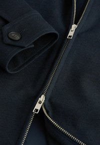 Marineblaues Zip-Up-Jackett mit glatter Textur, ausgestattet mit einem Metall-Reißverschluss, geknöpften Bündchen und klaren Linien im Stoffdesign.