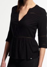 Blouse noire à boutons avec un décolleté en V, des manches trois-quarts et des plis horizontaux. Présente une taille froncée et des accents en dentelle.