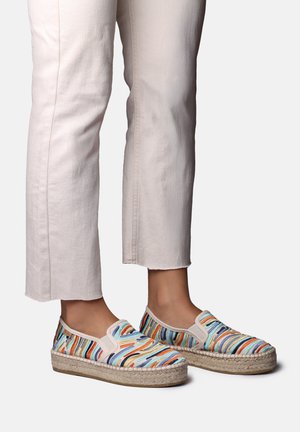 Nohy v nazúvacích espadrilách s viacfarebnými prešívanými pruhmi a béžovými platformovými podrážkami, v kombinácii s ľahko béžovými skrátenými nohavicami.
