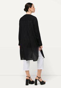 Schwarzes Langarmshirt mit einem transparenten Rückenteil, das ein strukturiertes Muster aufweist. Kombination mit weißen, verkürzten Hosen und klobigen schwarzen Absätzen.