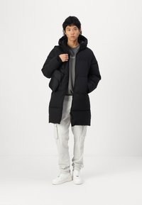 Abrigo acolchado negro con capucha, que presenta un cierre frontal, dos bolsillos laterales y un ajuste ligeramente oversize. Lleva puesto sobre una sudadera gris y pantalones claros.
