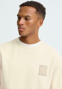 Beige sweatshirt met een ronde hals, voorzien van een gestructureerd, met tekst bedrukt patch. Zachte stof en een ontspannen pasvorm.