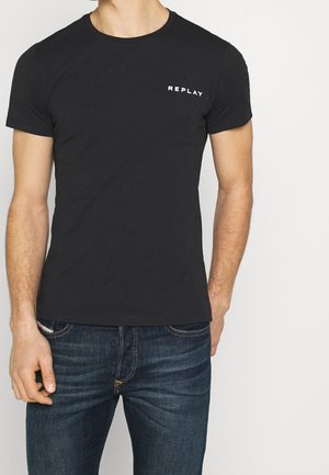 Mann trägt ein eng anliegendes schwarzes T-Shirt mit dem Text "REPLAY" auf der Brust und dunkelblaue Jeans vor einem einfarbigen hellen Hintergrund.