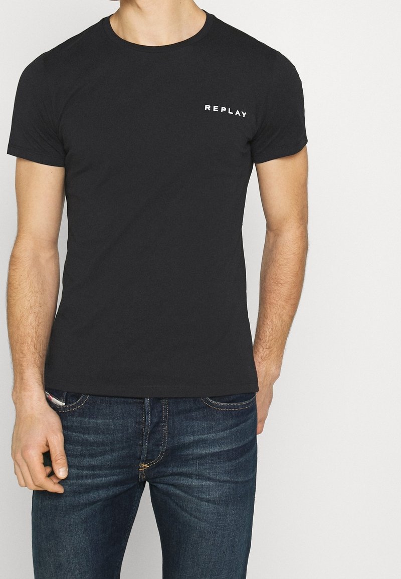 Homme portant un T-shirt noir ajusté avec le texte "REPLAY" sur la poitrine et un jean bleu foncé sur un fond clair uni.