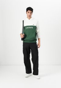 Roheline ja valge kapuutsiga sviiter "LACOSTE" logoga, millel on ees tasku; sobitatud mustade cargo-pükste ja valgete tossudega.