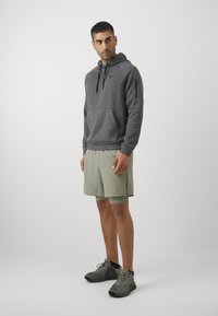 Pilkas hoodie su priekinėmis kišenėmis ir užtrauktuku, derinamas su šviesiai žaliais šortais ir juodais sportiniais batais. Vienoda fono spalva.