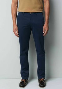 Navy blue tailored pants with a smooth texture, front pockets, and a button closure. Paired with black shoes featuring brown laces.