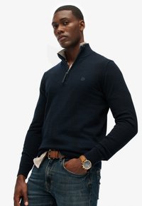 Marineblauwe pullover trui met een ritssluiting bij de kraag, geribbelde mouwen en een klein logo op de borst, gecombineerd met blauwe spijkerbroek en een bruine riem.