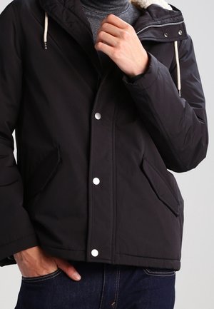 Schwarze isolierte Jacke mit Kapuze, ausgestattet mit einem Reißverschluss, Druckknöpfen und seitlichen Taschen. Weiche Fütterung an der Kragen sichtbar.