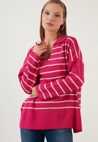 Roze prugasti pulover s V-izrezom, dugim rukavima i bijelim horizontalnim prugama. Tkanina izgleda mekano s opuštenim krojem i bočnim prorezima.