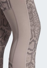 Detailní záběr na legíny s taupe hadím potiskem a hladkými, texturovanými panely látky podél strany nohy.