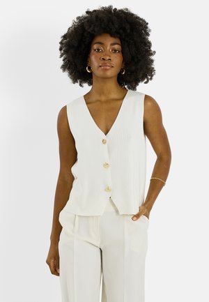 Femme aux cheveux naturellement bouclés porte un gilet blanc sans manches boutonné et un pantalon large assorti, accessoirisés de boucles d'oreilles anneaux en or et d'un bracelet.