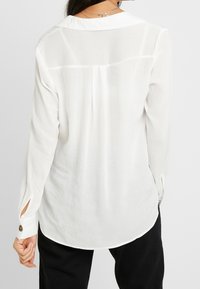 Camicia bianca a maniche lunghe con piega sul retro, orlo arrotondato e spacchi laterali. Dettagli con bottoni sui polsini e una texture morbida e leggera.