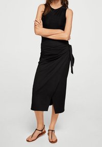 Schwarzes ärmelloses Wickelkleid mit taillierter Silhouette und seitlichem Bindegürtel. Sanfte Textur, knielang und mit einem subtilen Schlitz. Getragen mit flachen Sandalen.