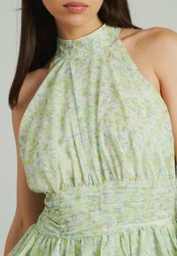 Jasnozielona sukienka w kwiaty z wysokim dekoltem, marszczonym stanem i zwiewnym dołem z delikatnymi plisami oraz subtelnym wzorem.