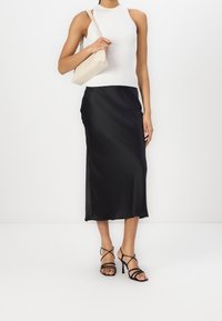 Mujer vistiendo una blusa sin mangas blanca, una falda midi negra, unos tacones altos negros con tiras y llevando un bolso de hombro beige.