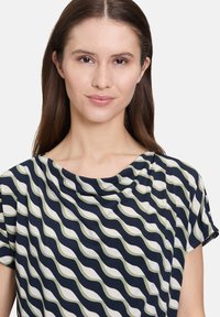 Krótki rękaw w granatowym kolorze z falistym wzorem w kremowym i zielonym odcieniu, z delikatnie podwiniętym dekoltem i gładkim, lekkim materiałem.