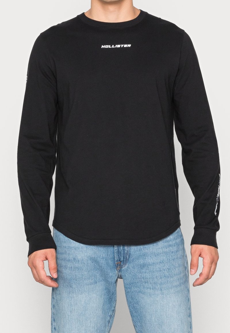 T-shirt noir à manches longues en coton, avec un col arrondi et un logo de la marque blanc sur la poitrine. Texture douce.