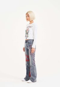 Camisa blanca de manga larga con un diseño gráfico colorido, combinada con jeans de denim acampanados decorados con bordados en rojo y azul.