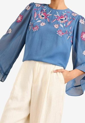 Blouse bleue transparente avec broderie florale rose et blanche sur la poitrine et les manches, portée avec un pantalon blanc taille haute et une main dans la poche.