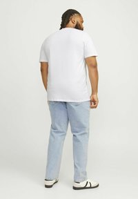 Camiseta blanca de manga corta hecha de algodón, combinada con jeans de mezclilla azul claro. Hombre visto desde atrás, de pie contra un fondo sencillo.