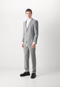 Completo su misura grigio chiaro con un sottile motivo a quadri, abbinato a una camicia bianca e scarpe nere stringate. Tessuto liscio, design aderente.