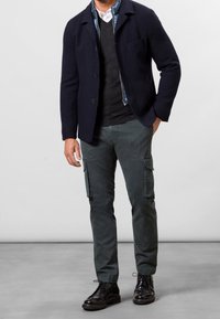 Námořnická texturovaná bunda přes šedý svetr, v kombinaci s tmavě zelenými cargo kalhotami a černými šněrovacími botami. Obsahuje podrobné švy a kapsy.