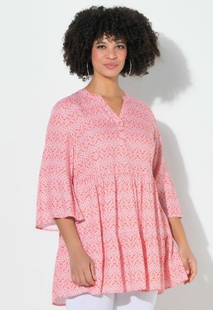 Kobieta o kręconych włosach, nosząca kolczyki koła i luźną, różową tunikę w wzory z rękawami długości 3/4, stojąca na tle białej ściany.