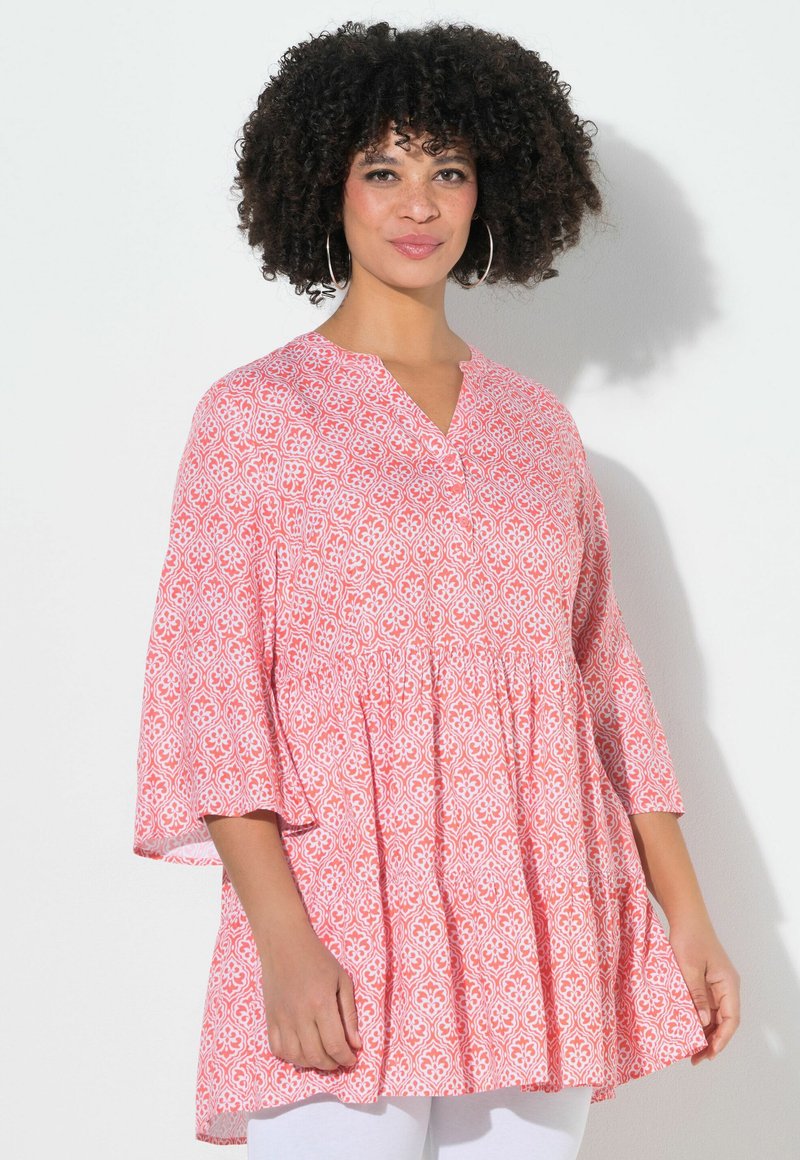 Vrouw met krullend haar, ringenoorbellen en een losse roze tuniek met patroon en driekwart mouwen, staand tegen een witte muur.