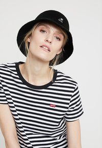Maglietta a righe bianco e nera con scollo rotondo e maniche corte, con un piccolo logo rosso di Levi's. Indossata con un cappello a secchio nero.