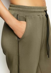 Oliwkowe dresy z elastycznym pasem, sznurkiem do regulacji i kieszeniami bocznymi. Tkanina jest gładka z kontrastującymi czarnymi detalami stębnowania.