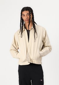Beige striped jacket with a zip-front, long sleeves, and side pockets. Features a classic collar and a fitted waistband.