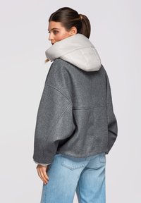 Femme aux cheveux bruns attachés en queue de cheval, portant une veste grise surdimensionnée avec une capuche rembourrée gris clair et un jean en denim bleu clair.