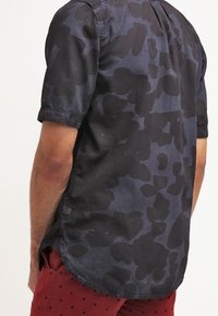 Mężczyzna ubrany w ciemnoniebieską koszulkę z krótkim rękawem z czarnym, abstrakcyjnym wzorem oraz czerwone szorty w małe czarne kropki, widziany od tyłu.