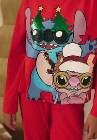 Il set di pigiama rosso presenta un design grafico con due personaggi che indossano accessori a tema natalizio, tra cui occhiali con paillettes e accenti festivi.